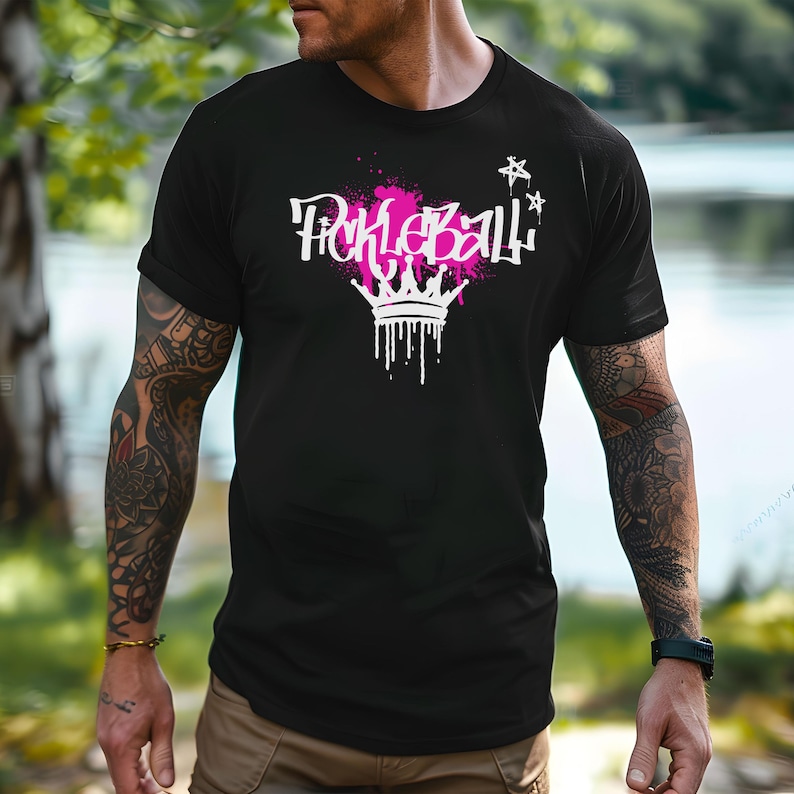 May include: Black t-shirt with a white and pink graphic that says "Pickleball" with a crown design. The text has a graffiti-style font with a pink paint splatter effect. Two white stars are above the text.