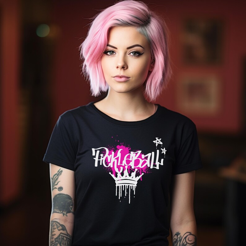 May include: Black t-shirt with the word "Pickleball" in white graffiti-style font, accented with pink paint splatters and a crown design. Two white stars are on the right side of the shirt.