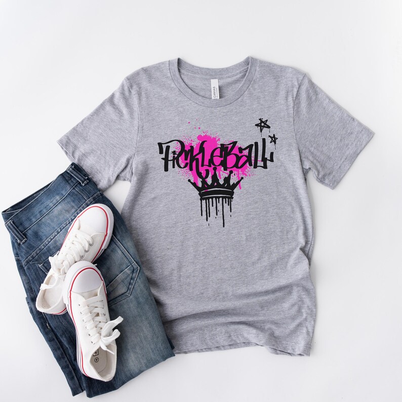 May include: A gray t-shirt with the word "Pickleball" in black graffiti-style lettering, a pink splatter design, and a black crown graphic. The shirt is paired with blue jeans and white sneakers.