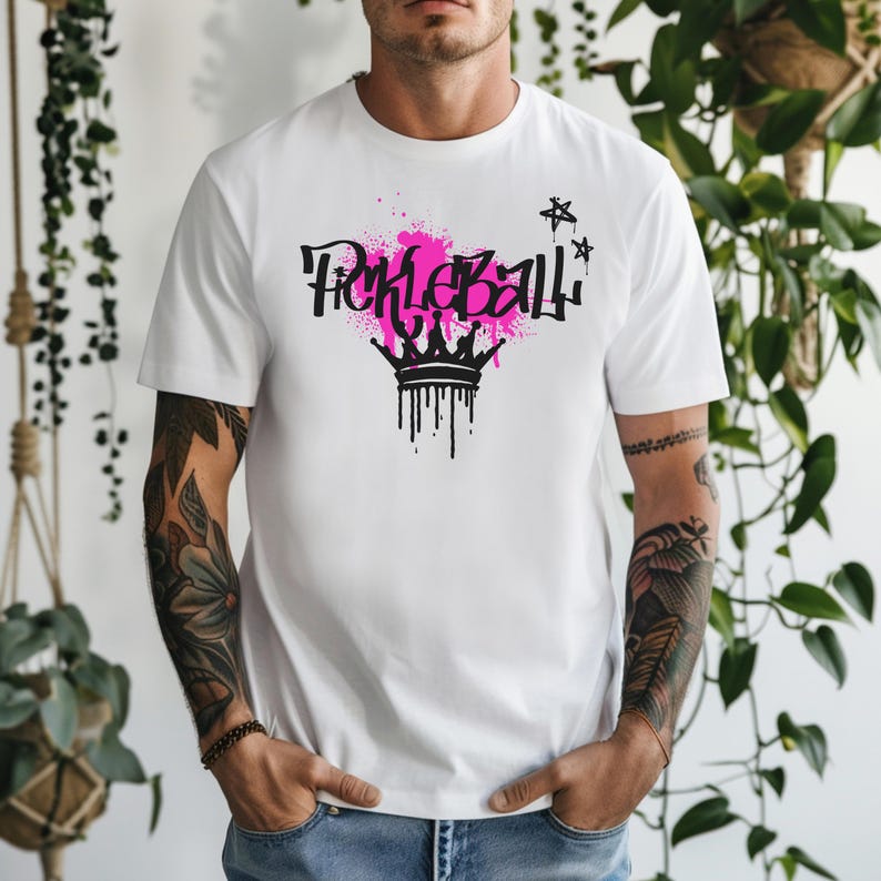 May include: White t-shirt featuring a graphic design with the word "Pickleball" in black graffiti-style lettering, set against a pink paint splatter. A black crown with dripping paint is below the text, with black stars on the right.