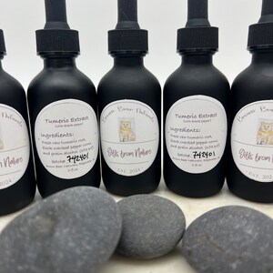 May include: Five black glass bottles with dropper tops, labelled "Brown Bear Naturals" and "Gifts from Nature". Each bottle contains a liquid with a label that reads "Tumeric Extract (with black pepper)" and lists the ingredients and batch number. The bottles are arranged in a row on a white surface with three grey stones in front of them.