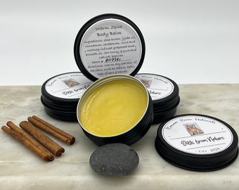 May include: A close-up of a small tin container filled with a yellow balm. The label reads "Warm Spice Body Balm" and lists the ingredients. The container is surrounded by cinnamon sticks and a small grey stone.