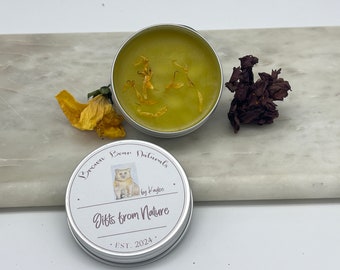 May include: A small, round, silver tin of green balm with dried flower petals. The lid features a bear illustration and the text "Brown Bear Naturals, Gifts from Nature, EST. 2024". A dried yellow flower and a dried red flower are also present.
