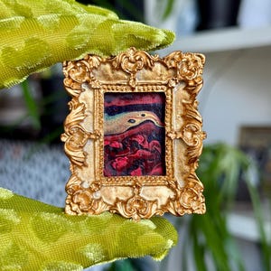 May include: A miniature gold-colored ornate picture frame with a small painting inside. The painting features red, blue, and yellow colors. The frame is held by a green, textured fabric.