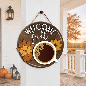 May include: A round wooden sign with the words "WELCOME fall" in white script. The sign features a coffee cup graphic and autumn leaves. The sign is hanging on a white wall, with pumpkins and lanterns in the background.