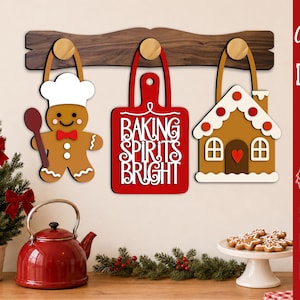 Puede incluir: Un estante de pared de madera oscura con tres ganchos redondos que sostienen decoraciones colgantes: una figura de pan de jengibre con gorro de chef y cuchara, una tabla de cortar roja con el texto BAKING SPIRITS BRIGHT y una casa de pan de jengibre.