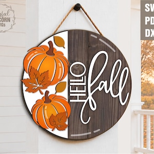 Könnte beinhalten: Runder Holzschild mit Herbstmotiv, mit orangefarbenen Kürbissen und Blättern. Das Schild zeigt "Hello Fall" in weißer Schrift. Es wird von einem braunen Seil gehalten. Der Hintergrund ist eine weiße Wand.