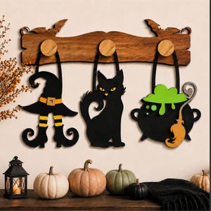 Puede incluir: Un perchero de madera con tres decoraciones colgantes: un sombrero de bruja negro con rayas naranjas, un gato negro con ojos amarillos y un caldero negro con líquido verde burbujeante y una escoba naranja, colgados de perillas redondas.