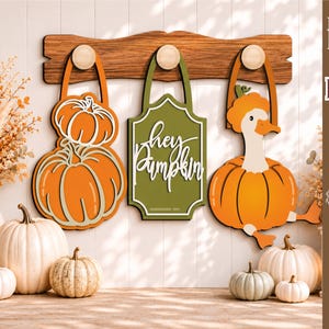 Könnte beinhalten: Eine hölzerne Wandgarderobe mit drei Haken, an denen hängende Dekorationen befestigt sind: zwei orangefarbene Kürbisse, ein grünes Schild mit dem weißen Text "hey Pumpkin" und eine weiße Gans in einem orangefarbenen Kürbiskostüm.