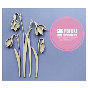 Op de afbeelding: Houten sneeuwklokjesbloemen, lasergesneden met ingewikkelde details, weergegeven tegen een lichtblauwe achtergrond. De bloemen hebben een natuurlijke houttint. Een roze cirkel bevat de tekst "SVG PDF DXF LASER CUT SNOWDROPS Joyful Unicorn SVGs."