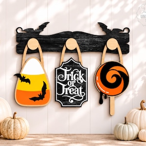 Puede incluir: Un perchero de pared de madera negra con siluetas de murci&eacute;lagos en los extremos y tres ganchos redondos. De los ganchos cuelgan una forma de caramelo de ma&iacute;z con murci&eacute;lagos, un letrero negro con texto blanco Trick or Treat y una piruleta.