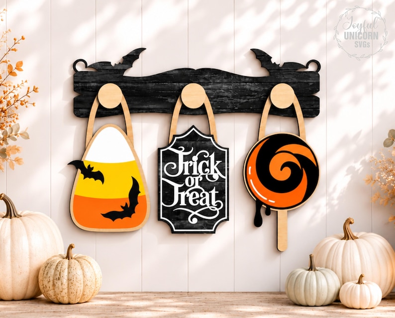 Puede incluir: Un perchero de pared de madera negra con siluetas de murci&eacute;lagos en los extremos y tres ganchos redondos. De los ganchos cuelgan una forma de caramelo de ma&iacute;z con murci&eacute;lagos, un letrero negro con texto blanco Trick or Treat y una piruleta.