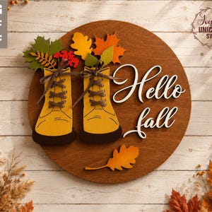 May include: A circular wooden wall sign featuring two yellow boots with dark brown soles and laces. The boots are decorated with colorful autumn leaves. The text Hello fall is written in white script. The sign is made of layered wood pieces.