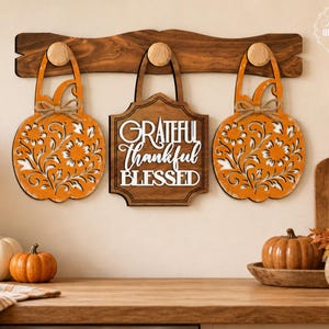 Puede incluir: Un perchero de madera oscura con tres ganchos redondos que sostienen dos recortes de madera naranja con forma de calabaza, patrones florales y lazos de cuerda, y una placa central de madera oscura con el texto blanco GRATEFUL THANKFUL BLESSED.