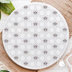 Peut inclure: Un objet décoratif rond en bois blanc avec un motif en nid d'abeilles et des abeilles. Les abeilles et le nid d'abeilles sont soulignés en gris foncé. Le motif est centré sur la surface en bois.