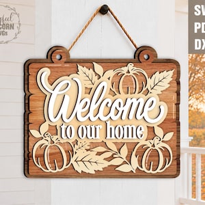 May include: A rectangular wooden hanging sign with a dark brown wood grain finish. The sign features layered light-colored cutouts of three pumpkins and autumn leaves. The text Welcome to our home is displayed in a white script font.