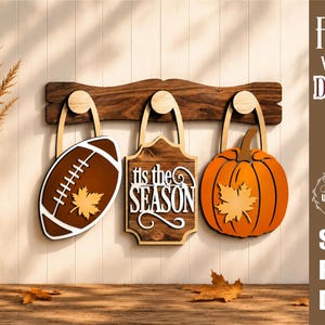 Op de afbeelding: Een houten wandrek met drie haken waaraan hangende decoraties zijn bevestigd: een bruine football met een esdoornblad, een rechthoekig bord met de tekst 'tis the SEASON' en een oranje pompoen met een esdoornblad.
