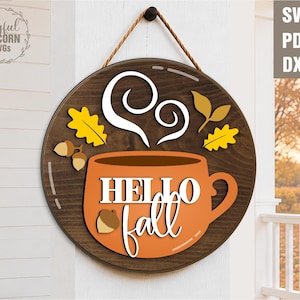 May include: A round wooden sign with a dark brown border and a rope hanger. The sign features an orange coffee mug with the words "HELLO fall" and autumn-themed accents like leaves and acorns. The sign also has the text "SVG PDF DXF".