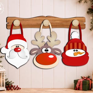 Puede incluir: Un perchero de pared de madera con tres ganchos, cada uno con un adorno navideño colgante. Los adornos muestran caras de estilo caricatura de Santa Claus, un reno con nariz roja y un muñeco de nieve con un gorro de invierno rojo.