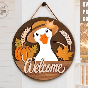 Può includere: Un cartello rotondo in legno raffigurante un'oca bianca che indossa un cappello marrone con una foglia arancione. Il design include una zucca arancione, spighe di grano e foglie d'acero. La parola Welcome è scritta in corsivo chiaro in basso.