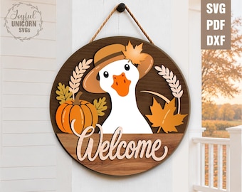 Op de afbeelding: Een rond houten bord met een witte gans die een bruine hoed met een oranje blad draagt. Het ontwerp bevat een oranje pompoen, korenaren en esdoornbladeren. Het woord Welcome staat onderaan in een lichtgekleurd schrijflettertype.