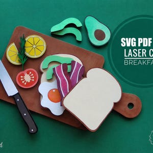 May include: A wooden cutting board with a variety of laser-cut breakfast food items. Includes a knife, a slice of lemon, tomato, avocado, bacon, egg, and a slice of toast. The text "SVG PDF DXF LASER CUT BREAKFAST" is visible.