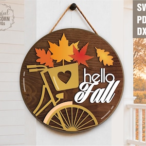 May include: A circular wooden wall sign featuring a dark brown stained finish and a hanging rope. The design includes a yellow bicycle with a heart-shaped basket, orange and red maple leaves, and the white text "hello Fall" in script font.