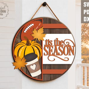 May include: A round wooden sign with a fall theme. It features a football, pumpkin, coffee cup, and fall leaves. The sign reads "It's the SEASON" in orange lettering. The sign is hanging by a brown rope.