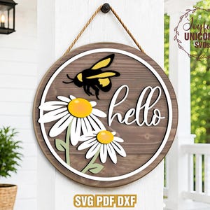May include: Circular wooden sign with a daisy and bee design. The sign has a white border, the word "hello" in cursive, and the text "SVG PDF DXF" at the bottom. It is suspended by a rope.