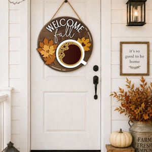 May include: A round wooden sign with the words "WELCOME fall" and a coffee cup design. The sign is decorated with fall leaves and hangs on a white door. A welcome mat and pumpkins are in front of the door.