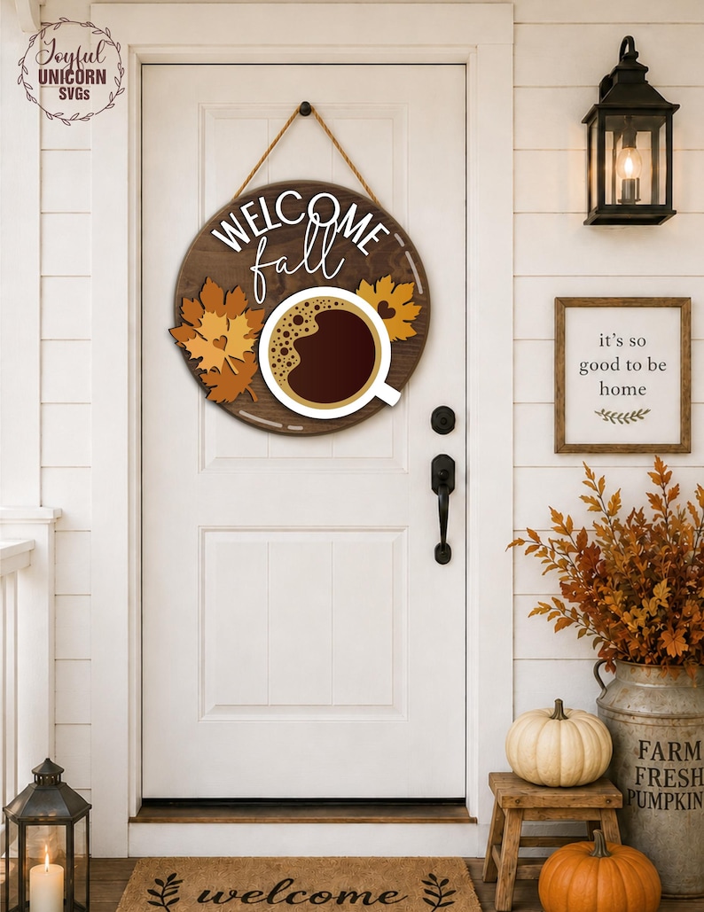 May include: A round wooden sign with the words "WELCOME fall" and a coffee cup design. The sign is decorated with fall leaves and hangs on a white door. A welcome mat and pumpkins are in front of the door.