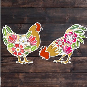 May include: Two-piece layered SVG, PDF, and DXF cut file set featuring a hen and rooster. The hen and rooster are decorated with floral designs in shades of red, pink, white, and green. The background is a dark wood grain.