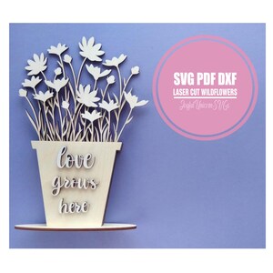 May include: A wooden laser-cut flower pot with the words "love grows here." The pot holds white daisy-like flowers. The image includes the text "SVG PDF DXF Laser Cut Wildflowers" in a pink circle.