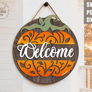 May include: A round wooden welcome sign in the shape of a pumpkin. The sign features orange and green pumpkin details with the word "Welcome" in white script. It hangs from a brown rope.