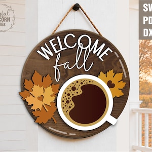 May include: A round wooden sign with the words "WELCOME fall" in white script. The sign features a coffee cup, fall leaves, and a rope for hanging. The sign is a fall-themed decoration.