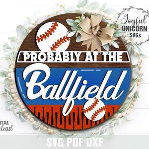以下が含まれることがあります： 「PROBABLY AT THE Ballfield」の白い文字が入った丸い木製の看板。赤、白、青の野球ボールとバットのデザインが特徴です。装飾用のリボンと緑が看板を囲んでいます。