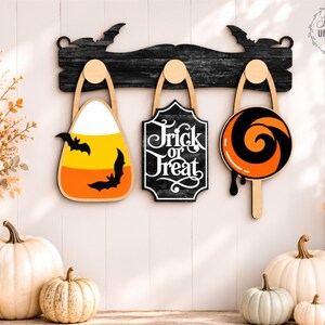 Puede incluir: Un estante de pared de madera negra con siluetas de murci&eacute;lagos sostiene tres decoraciones colgantes: una forma de caramelo de ma&iacute;z con dos murci&eacute;lagos, un letrero negro con texto blanco Trick or Treat y una piruleta naranja y negra.