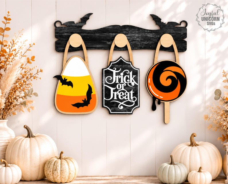 Puede incluir: Un estante de pared de madera negra con siluetas de murci&eacute;lagos sostiene tres decoraciones colgantes: una forma de caramelo de ma&iacute;z con dos murci&eacute;lagos, un letrero negro con texto blanco Trick or Treat y una piruleta naranja y negra.