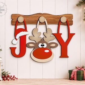 Puede incluir: Un letrero de pared de madera con la palabra JOY colgada de un tablero horizontal. La letra J es roja con un gorro de Papá Noel con borde blanco, la letra O es reemplazada por una cara de reno marrón con nariz roja y la letra Y es roja.