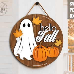 May include: A round wooden sign featuring a white ghost, two orange pumpkins, and yellow maple leaves. The text "hello Fall" is written in white script and sans-serif font. The sign hangs from a brown rope and includes SVG, PDF, and DXF labels.