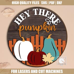 Op de afbeelding: Een rond houten bord met de tekst "HEY THERE pumpkin" in wit en oranje. Het bord heeft een ontwerp van witte, rode en blauwgroene pompoenen met een herfstblad en een hek. Voor lasers en snijmachines.
