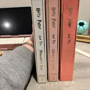 May include: Three hardcover books from the Harry Potter series. The spines are white, pink, and coral, with gold lettering. The titles are "Harry Potter and the Philosopher's Stone," "Chamber of Secrets," and "Prisoner of Azkaban."