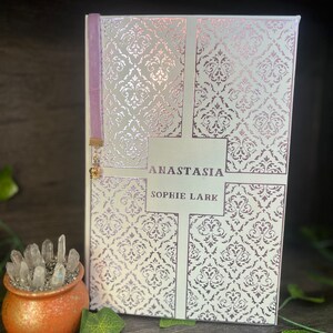 May include: A white book with a silver and pink damask pattern on the cover. The title "Anastasia" and the author "Sophie Lark" are printed in black on the front cover. A purple ribbon bookmark is attached to the spine.