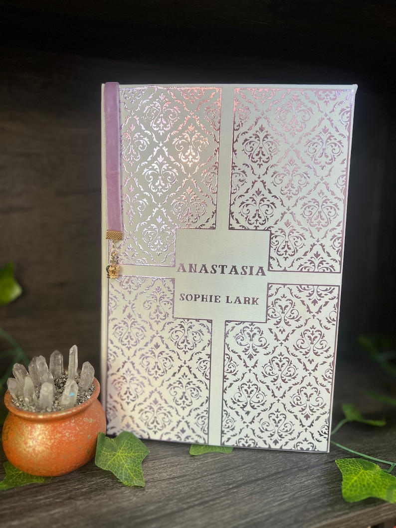 May include: A white book with a silver and pink damask pattern on the cover. The title "Anastasia" and the author "Sophie Lark" are printed in black on the front cover. A purple ribbon bookmark is attached to the spine.