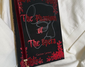 May include: A hardcover book titled "The Phantom of the Opera" by Gaston Leroux. The black cover features a mask design and red floral accents. A red velvet bookmark with a gold charm is inserted.