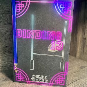 May include: A black book with a pink and blue border. The title "BINDING 13" is in a pink and blue gradient font. The author "CHLOE WALSH" is printed in white below the title. A blue ribbon bookmark with a gold star charm is attached to the book.
