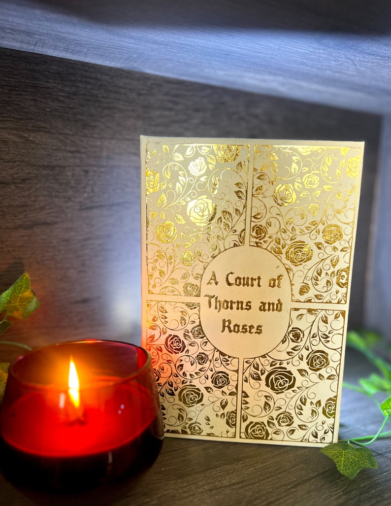 May include: A white book with a gold foil design of roses and vines. The title "A Court of Thorns and Roses" is printed in gold on the front cover.