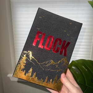May include: A black book with a gold and red design. The title "FLOCK" is printed in red on the front cover. The design features a mountain range and trees in gold foil.
