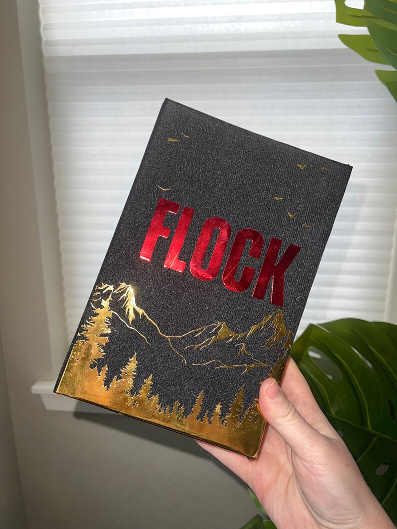 May include: A black book with a gold and red design. The title "FLOCK" is printed in red on the front cover. The design features a mountain range and trees in gold foil.