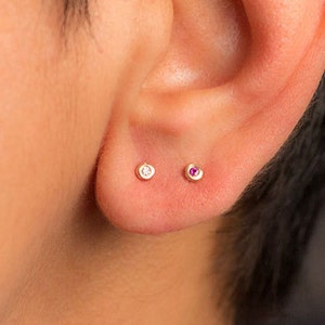 Może przedstawiać: Ucho z dwoma małymi kolczykami. Lewy kolczyk ma przezroczysty kamień, a prawy kolczyk ma r&oacute;żowy kamień.